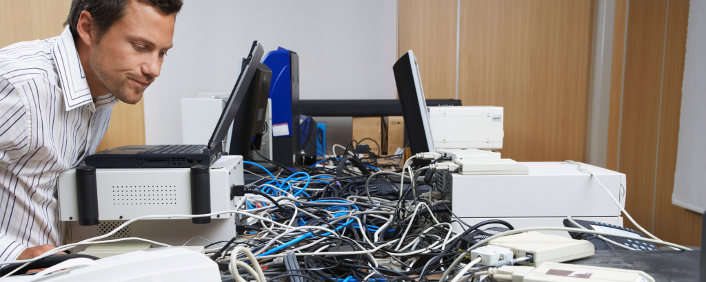 Stressed office worker surrounded by tangled cables and unmanaged tech equipment, representing the chaos of DIY IT support.